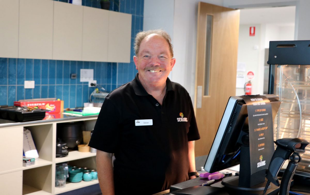 Man smiles working behind food service counter at Seasons Cafe