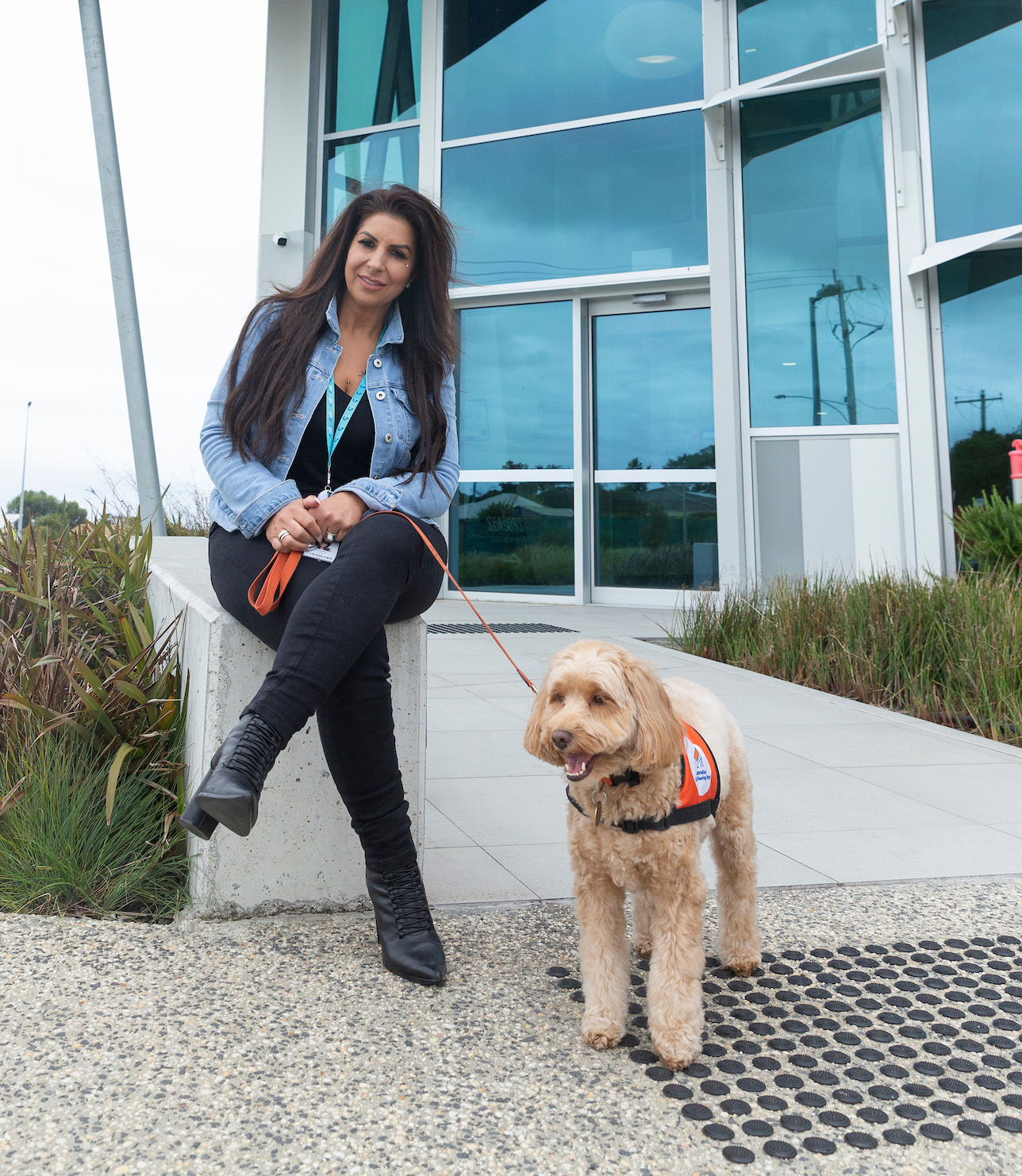A smiling Danielle sitting outside the genU Support Hub in Highton with her Lions hearing dog, Honey who is a gold labradoodle.