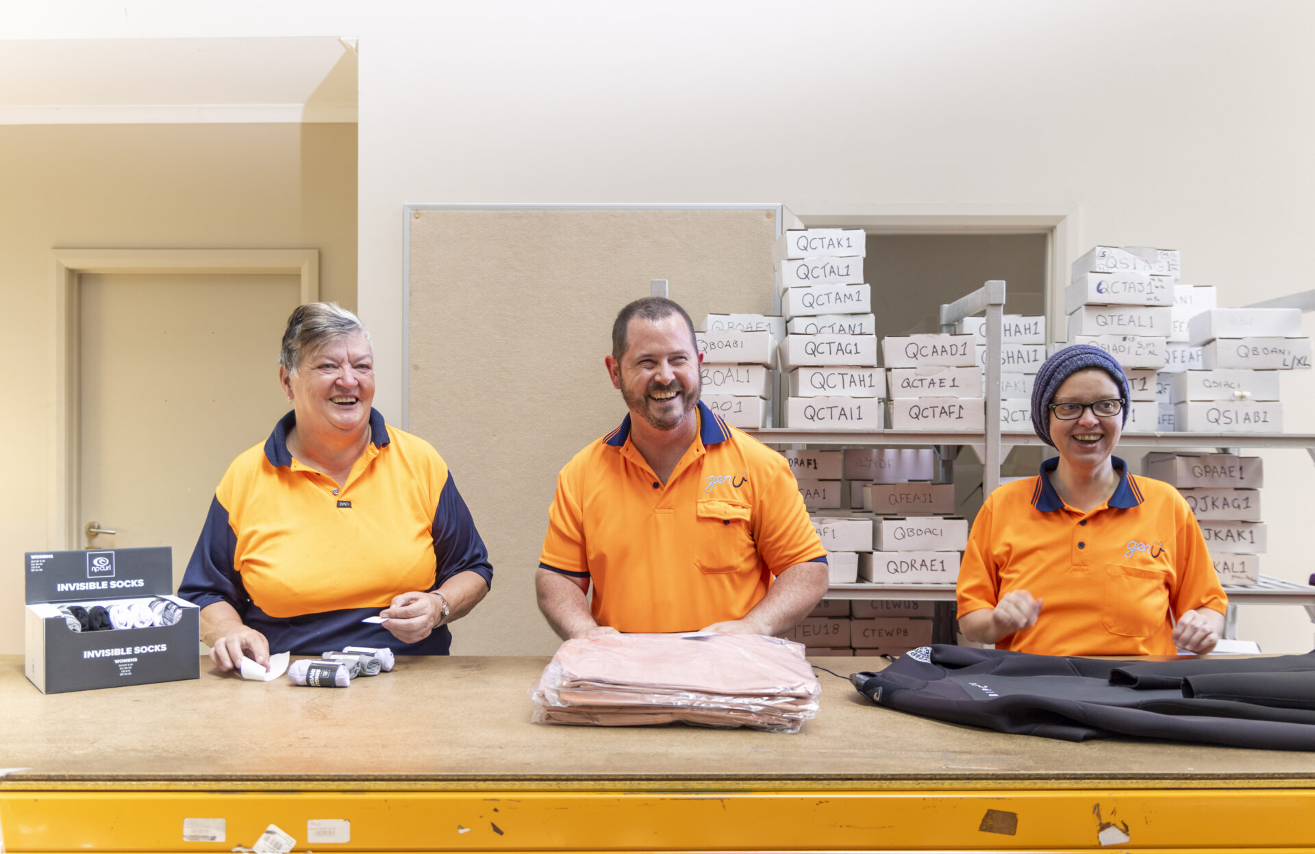 A smiling Andrew with two female supported colleagues who are also smiling at a warehouse facility packing Rip Curl wetsuits, socks and clothing.