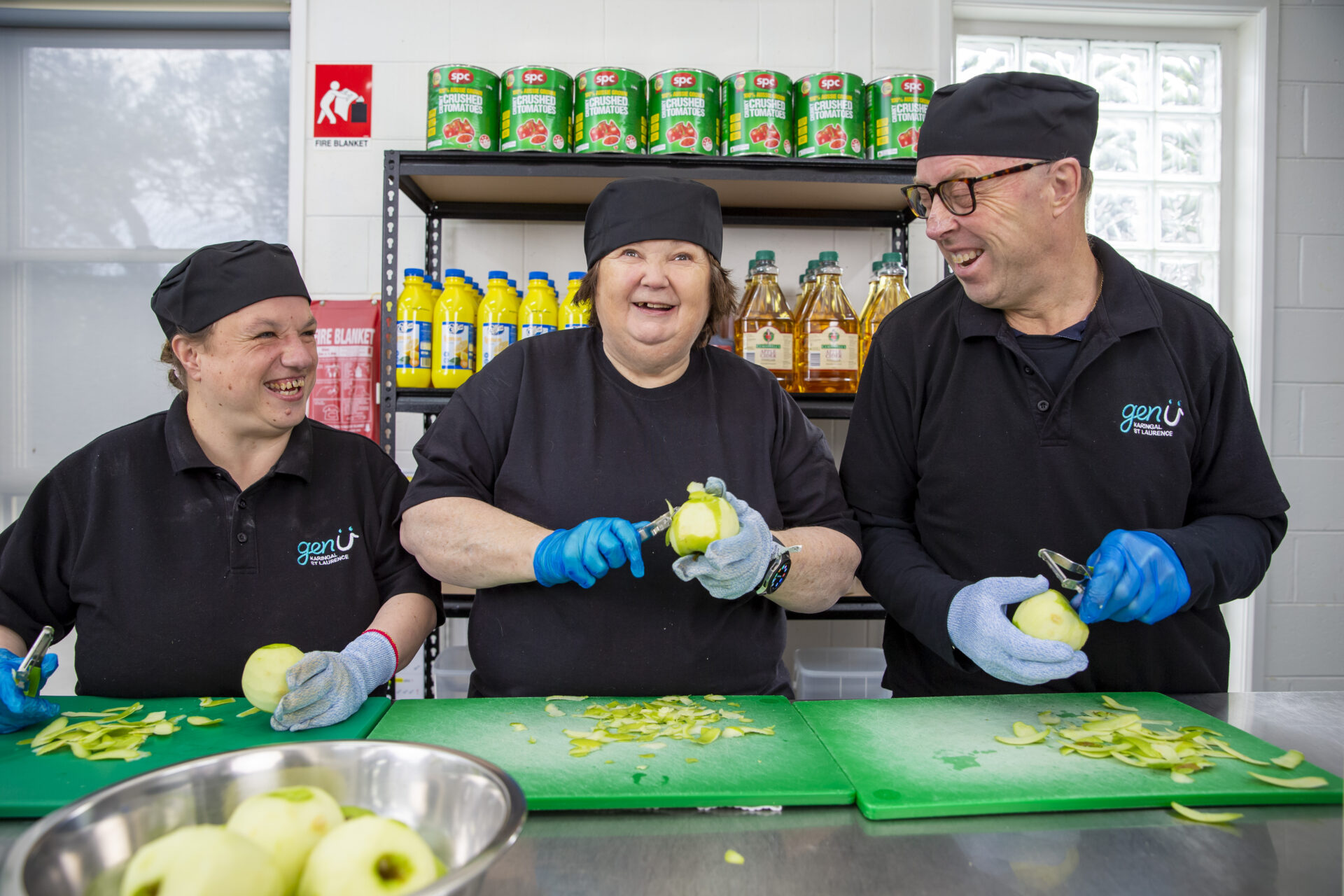 Kerrie is peeling apples with two other supported employees at the Seasons Kitchen in Rosebud. All supported employees are smiling and wearing black hats and blue gloves.