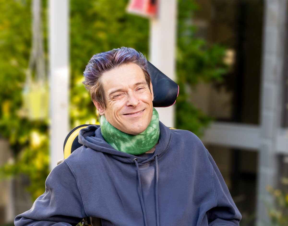 A smiling Patrick is seated in his mobility chair outside of Oakdale Lodge in Tasmania.