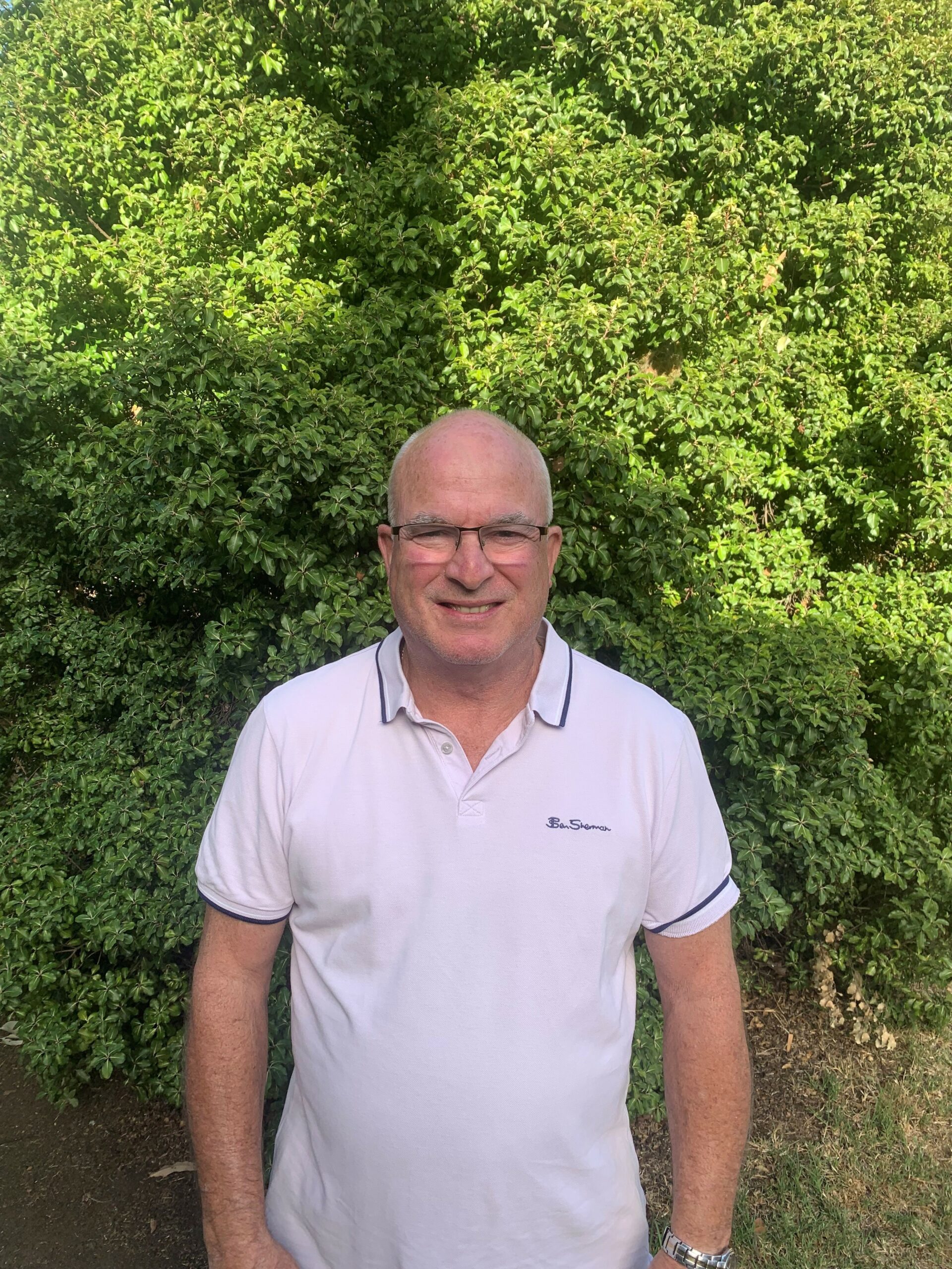 Greg is outside against a backdrop of green trees. he is smiling and wearing a white polo top and glasses