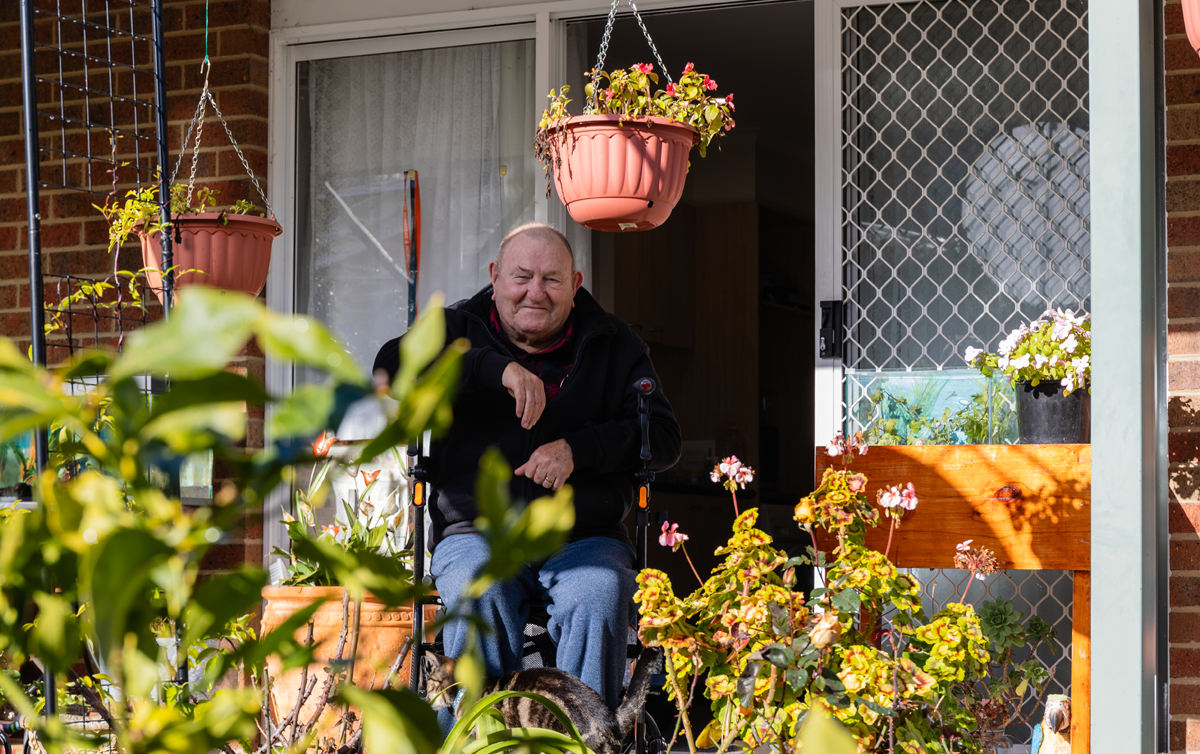 An elderly man is sitting on a walker on his patio. He is surrounded by a garden on a sunny day.