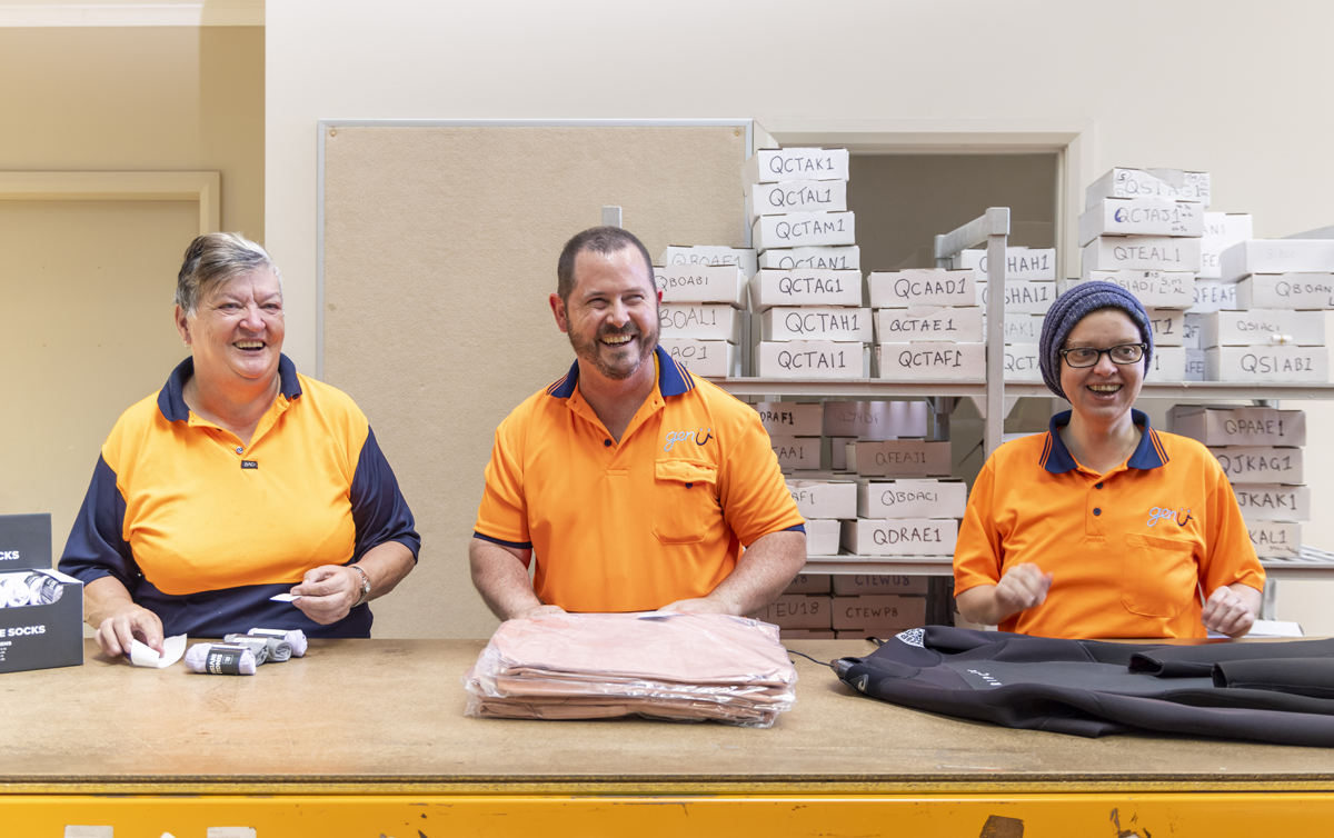 Three genU supported employees are standing at a table packing orders in a Rip Curl warehouse.