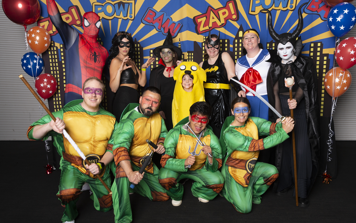 A group of people are wearing fancy dress costumes in front of a superhero backdrop for the genU Big Night Out.